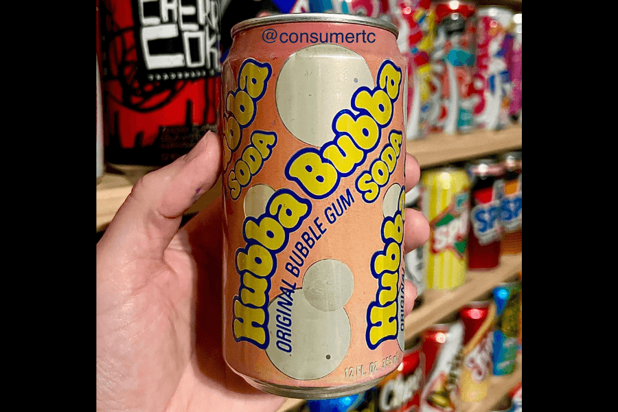 This image features a hand holding a can of Hubba Bubba Soda, a bubble gum-flavored drink with a retro design. The can has a peach-colored background with bold blue and yellow text and bubble illustrations, evoking a nostalgic feel.