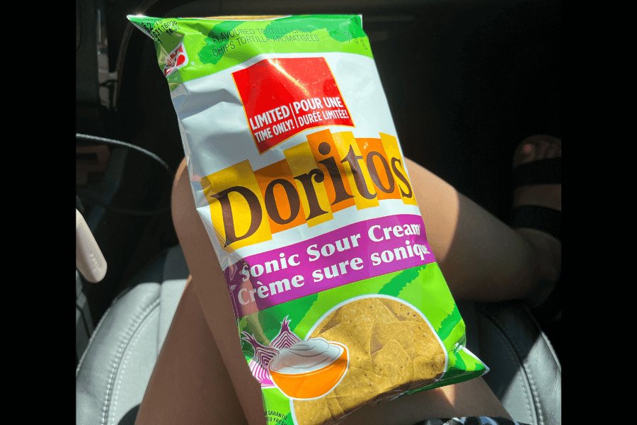 A bag of Doritos in the "Sonic Sour Cream" flavor with green and white packaging, labeled as a "Limited Time Only" release in English and French. The design features tortilla chips with a bowl of sour cream and onions at the bottom. The bag is being held on someone's lap inside a car, with sunlight highlighting the packaging.