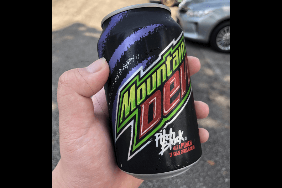 This image shows a hand holding a can of Mountain Dew Pitch Black, featuring a bold black design with purple accents and vibrant green and red branding. The label highlights the flavor as "a punch of grape citrus," standing out against the dark background.