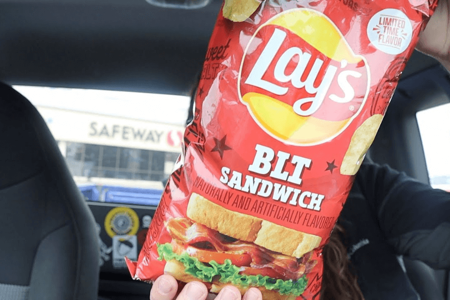 A bag of Lay's chips in the "BLT Sandwich" flavor with a vibrant red design. The packaging features an image of a BLT sandwich with crispy bacon, lettuce, and tomato between slices of bread, along with a "Limited Time Flavor" label. The bag is being held inside a car, with the Safeway store sign visible through the window in the background.