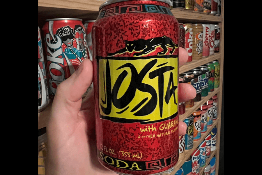 This image shows a hand holding a can of Josta soda, featuring a bold red background with a yellow label and black lettering. The design includes a black panther graphic and the text "with guarana & other natural flavors," giving it a unique, energetic vibe.