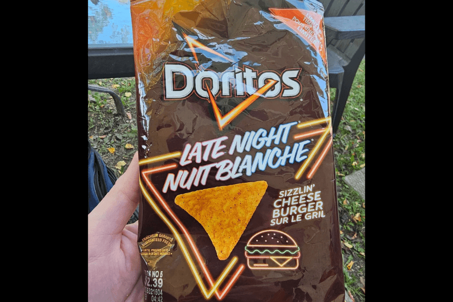 A bag of Doritos in the "Late Night Sizzlin' Cheese Burger" flavor with a dark brown design. The packaging features neon-style text reading "Late Night / Nuit Blanche" and an illustration of a cheeseburger with glowing accents. The bag is held outdoors, with a table and grass visible in the background.