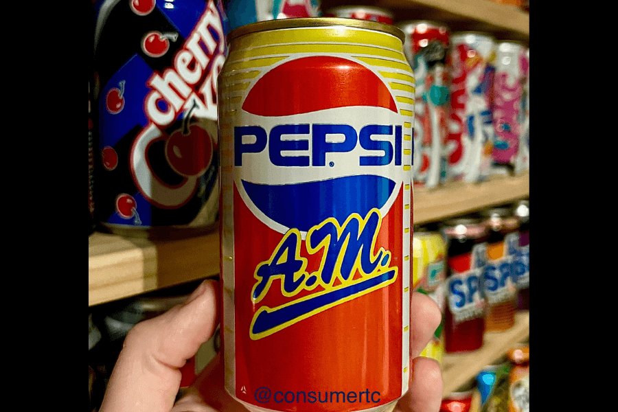 This image features a hand holding a can of Pepsi A.M., designed with a vibrant orange, blue, and yellow color scheme. The logo includes bold "A.M." lettering beneath the iconic Pepsi branding, suggesting a morning-themed beverage.