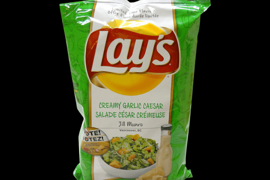 A bag of Lay's chips in the "Creamy Garlic Caesar" flavor, featuring a green and white design. The packaging includes an image of a Caesar salad with croutons and a bottle of Caesar dressing. It highlights a limited-time flavor and credits Jill Munro from Vancouver, BC, as the flavor's creator. A "Vote!" promotion is displayed near the bottom of the bag.