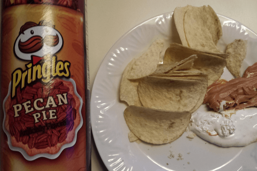 A can of Pringles in the "Pecan Pie" flavor with a brown design featuring pecan imagery on the label. Next to the can is a plate containing a serving of Pringles chips alongside dollops of marshmallow fluff and a spread of caramel or peanut butter. The setup is displayed on a light-colored tabletop.