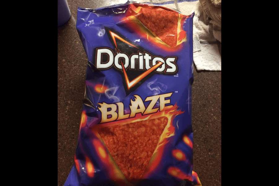 A bag of Doritos in the "Blaze" flavor with a bold purple design. The packaging features a fiery graphic surrounding the Doritos logo and a large triangular chip, emphasizing its intense spiciness. The bag is placed on a dark countertop, with a towel visible in the background.