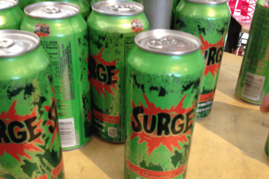 This image features several green cans of "SURGE," a citrus-flavored soda, displayed on a table or shelf. The cans are brightly decorated with bold black and orange lettering, splashes of green, and a retro, energetic design.