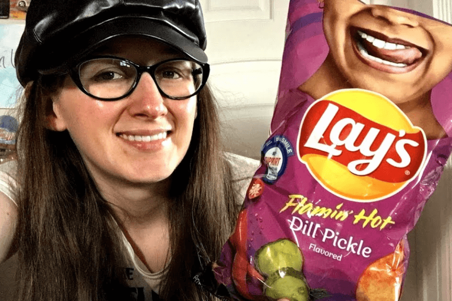 A woman wearing glasses and a black leather cap is smiling while holding a bag of Lay's chips in the "Flamin' Hot Dill Pickle" flavor. The packaging is purple with an image of pickles and chili peppers, along with a playful design of a person sticking out their tongue. The bag also features a "Support Our Future Farmers" emblem.