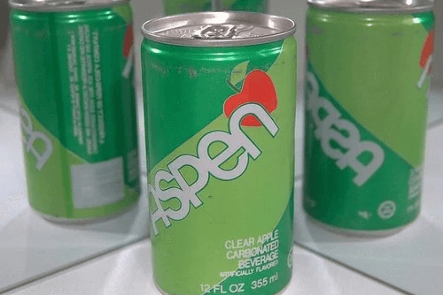 This image shows a can of Aspen soda, a clear apple-flavored carbonated beverage. The design features a green background with a red apple graphic and white text, emphasizing its crisp and fruity appeal.