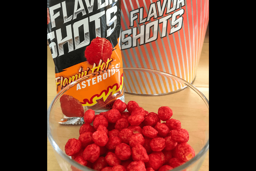 A package of Flamin' Hot Asteroids from the "Flavor Shots" series is displayed alongside a clear glass bowl filled with the bright red, spicy snack. The packaging features bold black and orange designs with flame graphics, emphasizing the fiery flavor. A striped "Flavor Shots" container is visible in the background.