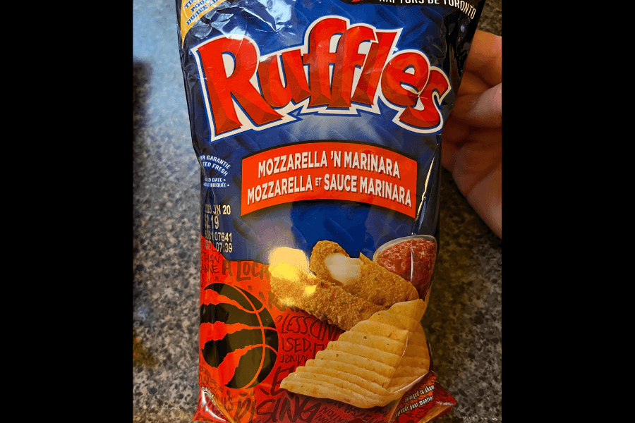 A bag of Ruffles chips in the "Mozzarella 'n Marinara" flavor, featuring a navy blue and red design. The packaging showcases an image of mozzarella sticks with marinara sauce alongside ridged chips. A Toronto Raptors basketball emblem and team branding are displayed at the bottom, indicating a promotional collaboration. The bag is held over a granite countertop.