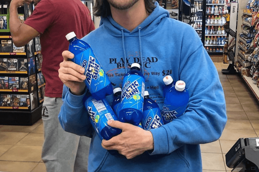 This image shows a person wearing a blue hoodie holding several bottles of "Mondo Arctic Sun," a blue beverage with lemon-lime flavor branding. The scene appears to be inside a convenience store, with shelves of snacks and products visible in the background.