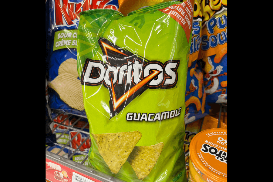 A bag of Doritos in the "Guacamole" flavor with a bright green design. The packaging features two triangular chips with visible green seasoning to emphasize the flavor. It is displayed on a store shelf alongside other snacks, including Ruffles Sour Cream and Cheddar chips and Cheetos Puffs.