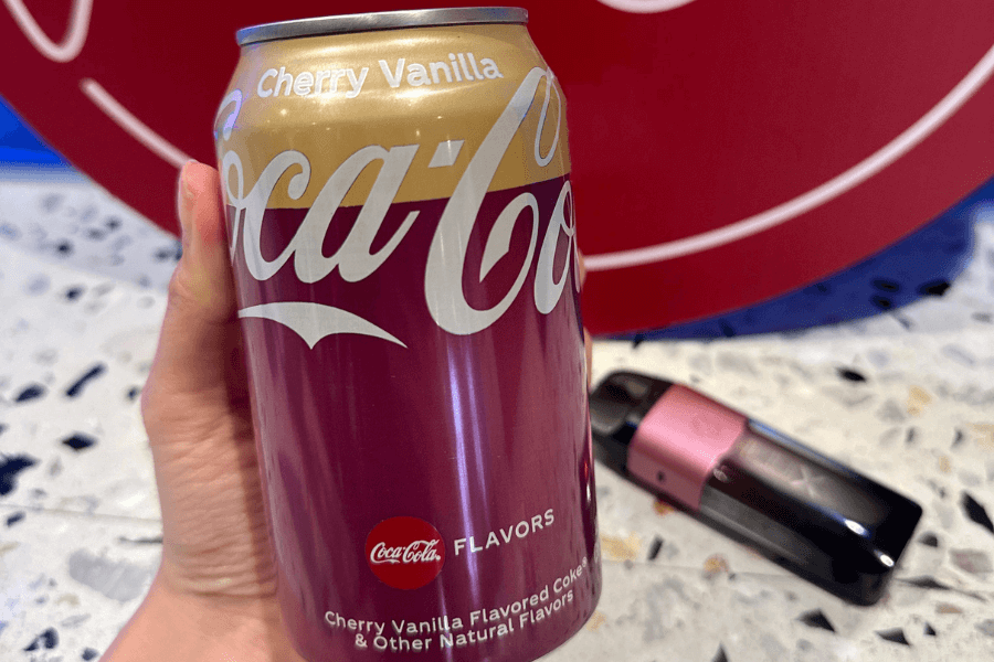 This image shows a can of Coca-Cola Cherry Vanilla, featuring a gradient design of gold and cherry red with bold white branding. The label highlights its unique cherry and vanilla flavor, and the can is held against a marble-like surface with a red Coca-Cola logo in the background.