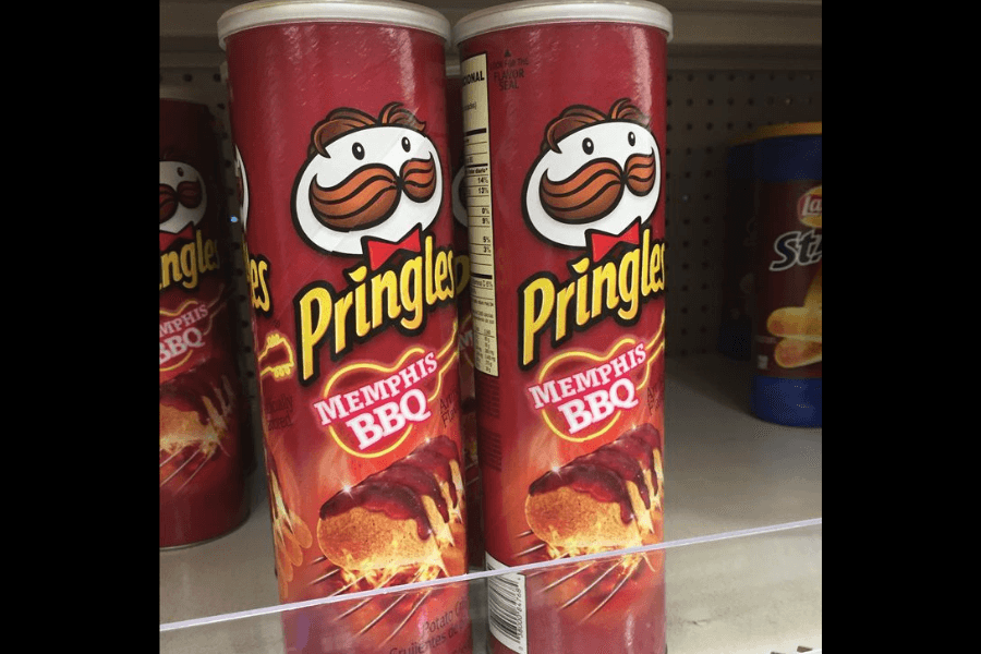 Cans of Pringles in the "Memphis BBQ" flavor with a red design prominently featuring a barbecue rib covered in sauce and flames in the background. The Pringles mascot is displayed at the top of the can. The cans are lined up on a store shelf, with other snack items partially visible in the background.