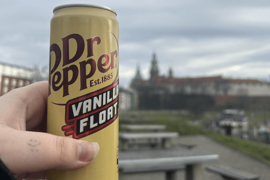 This image shows a hand holding a can of Dr Pepper Vanilla Float with a light yellow design and bold maroon branding. The background features an outdoor setting with blurred buildings and a scenic view, creating a casual and relaxed atmosphere.