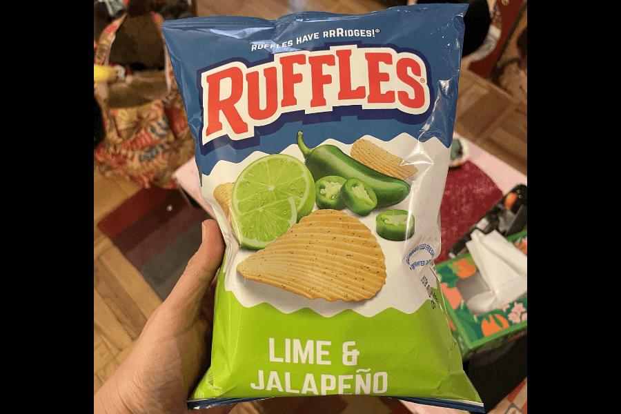 A bag of Ruffles chips in the "Lime & Jalapeño" flavor, featuring a green and blue design. The packaging showcases ridged chips alongside slices of lime and jalapeño peppers, emphasizing the tangy and spicy flavor combination. The bag is held indoors, with a colorful and busy background featuring household items.