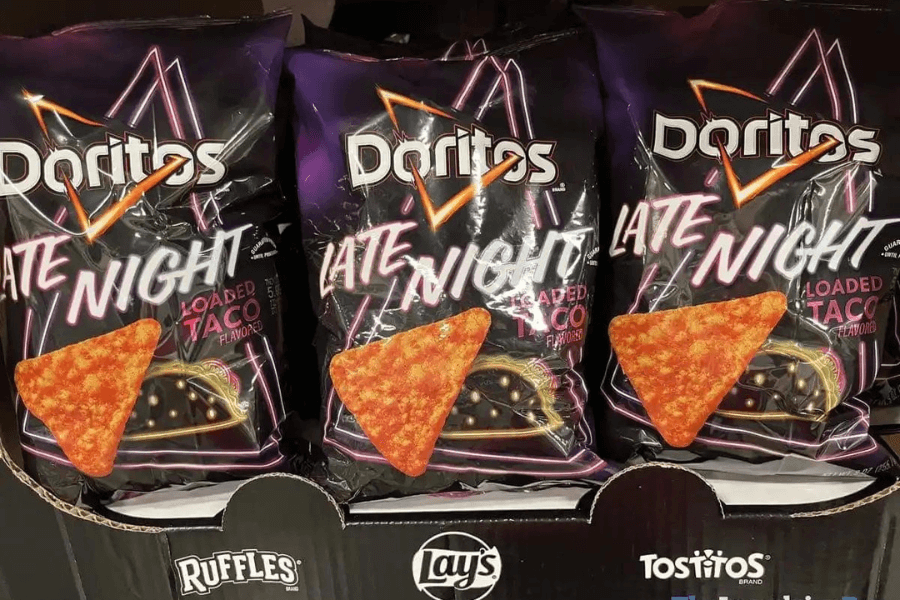 Bags of Doritos "Late Night Loaded Taco" flavor displayed on a store shelf, featuring a dark purple design with neon-style graphics. The packaging showcases a triangular Dorito chip and a loaded taco, emphasizing the bold flavor. The bags are arranged in a branded display alongside logos for Ruffles, Lay's, and Tostitos at the bottom.