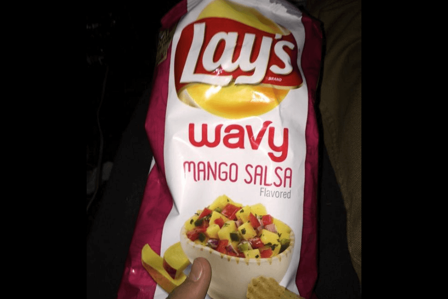 A bag of Lay's Wavy chips in the "Mango Salsa" flavor, featuring a white and pink design. The packaging displays a bowl of colorful mango salsa made with diced mangoes, red bell peppers, and herbs. A chip is being held in front of the bag, suggesting it pairs well with the salsa-inspired flavor.