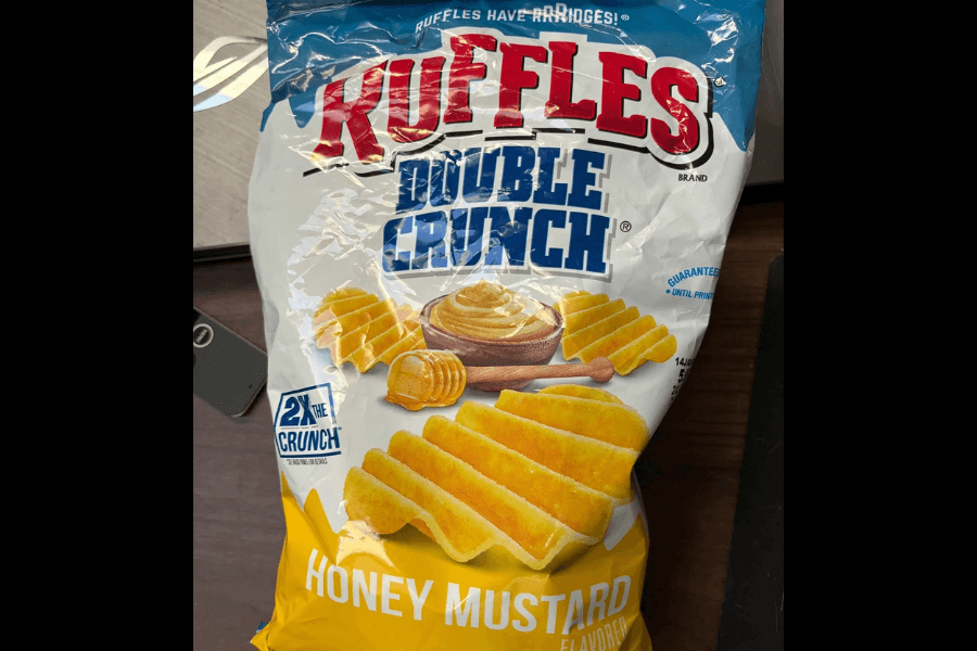 A bag of Ruffles Double Crunch chips in the "Honey Mustard" flavor, featuring a blue and white design with a yellow accent. The packaging displays the ridged, extra-crunchy chips alongside a bowl of honey mustard and a honey dipper. The label highlights "2X the Crunch" for its texture and boldness. The bag is resting on a desk with office items visible in the background.