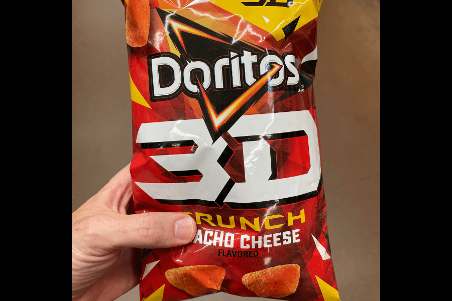 A bag of Doritos 3D Crunch chips in the "Nacho Cheese" flavor, featuring a bold red, white, and yellow design. The packaging highlights the 3D puffed texture of the chips with vibrant graphics and a prominent "3D" logo. A hand is holding the bag against a neutral indoor background.