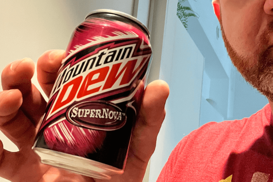 This image shows a person holding a can of Mountain Dew SuperNova, featuring a striking pink and black design with bold white and red branding. The label emphasizes its unique flavor and energetic appeal with bright, dynamic graphics.