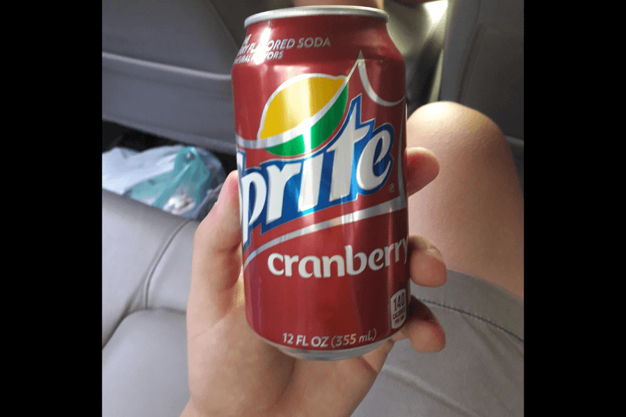 This image shows a hand holding a 12-ounce can of Sprite Cranberry, featuring a red design with the classic Sprite logo and cranberry-themed accents. The can emphasizes its seasonal cranberry flavor while maintaining Sprite's signature branding.