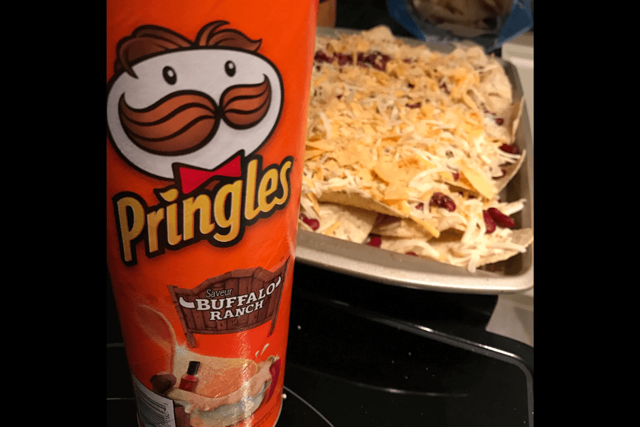 A can of Pringles in the "Buffalo Ranch" flavor with an orange design, featuring an image of dipping sauce and a wooden sign branding the flavor. In the background, a tray of nachos topped with shredded cheese and other ingredients is visible, suggesting a savory snack pairing. The setup is displayed on a kitchen countertop.