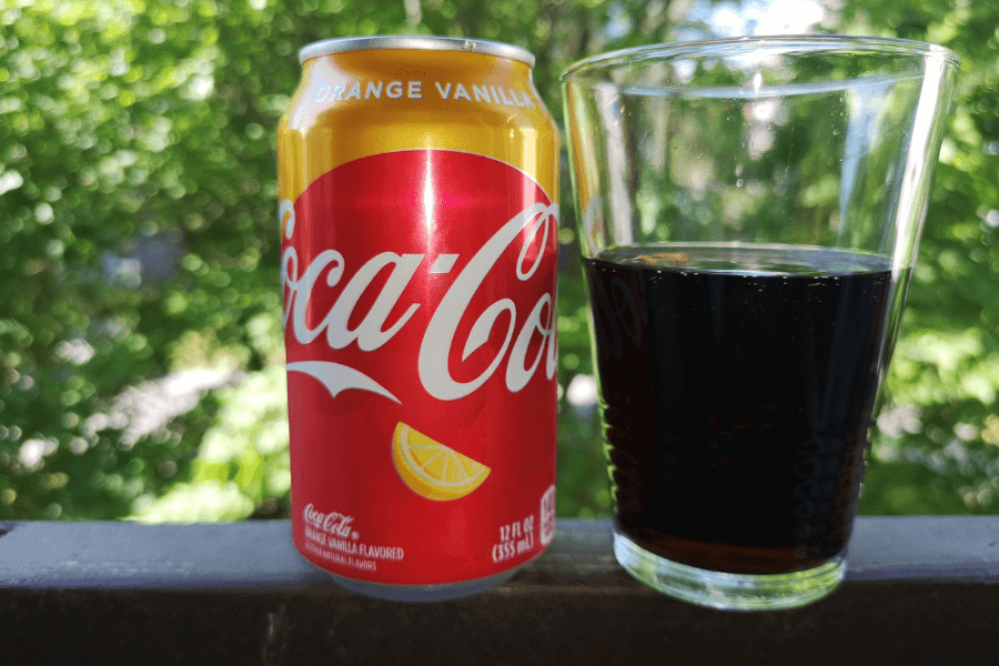 This image showcases a can of Coca-Cola Orange Vanilla alongside a glass filled with the soda. The can features a bright orange and red design with white text and a citrus slice graphic, emphasizing the unique orange and vanilla flavor. The background includes lush greenery, adding a fresh, outdoor vibe.