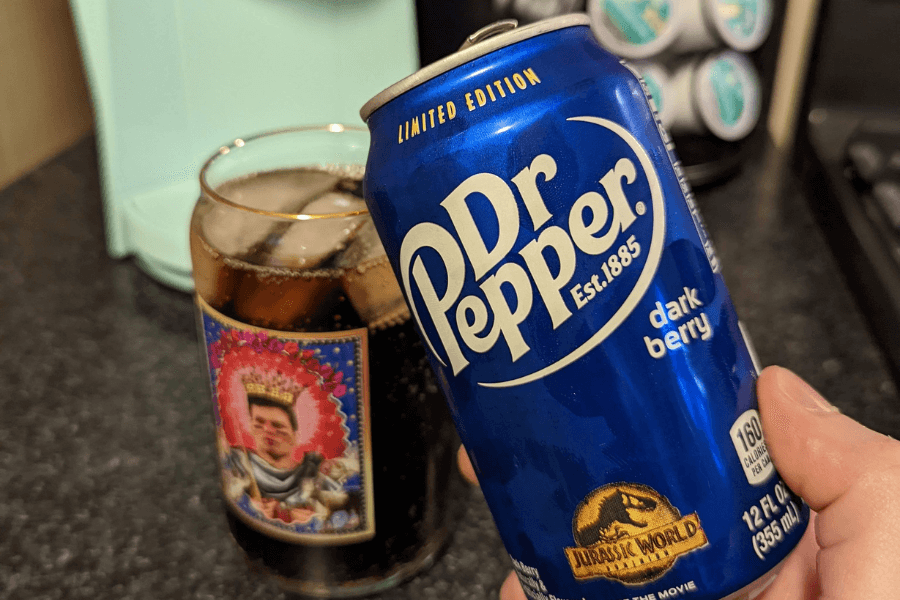 This image shows a limited edition can of Dr Pepper Dark Berry, featuring a deep blue design with bold white text and a Jurassic World logo at the bottom. In the background, a glass filled with the soda and ice is visible, adding to the promotional and refreshing appeal.