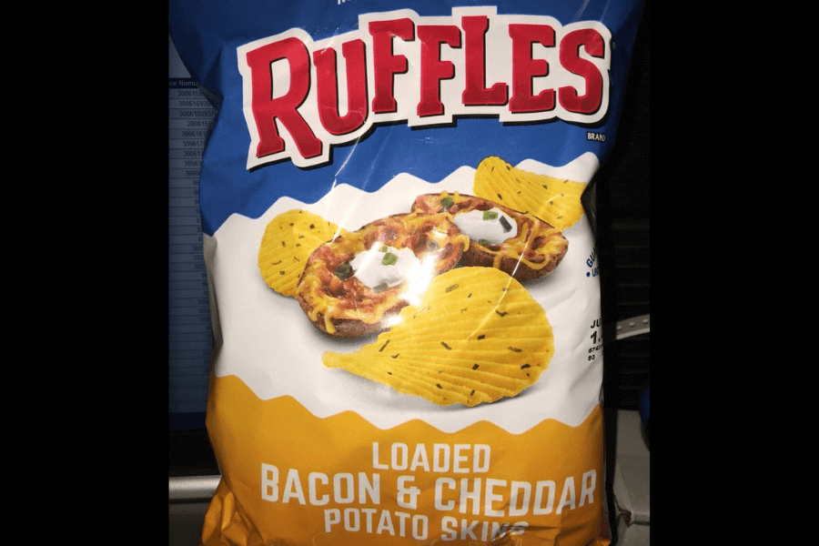 A bag of Ruffles chips in the "Loaded Bacon & Cheddar Potato Skins" flavor, featuring a blue and yellow design. The packaging displays ridged chips alongside a serving of loaded potato skins topped with cheese, bacon, sour cream, and chives, emphasizing the rich and savory flavor. The bag is photographed indoors with a computer screen faintly visible in the background.