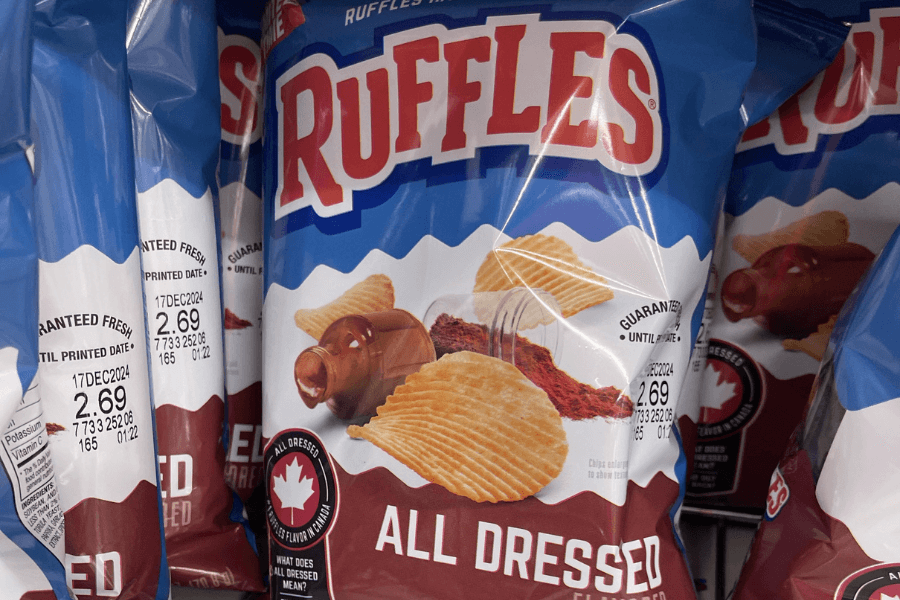 A bag of Ruffles potato chips in the "All Dressed" flavor, featuring a blue, red, and white design. The packaging displays a bottle of vinegar, spices, and ridged chips to highlight the flavor combination, along with a small Canadian flag emblem reading "All Dressed: Canada's Favorite Flavor." Multiple bags are shown on a store shelf, with price and freshness guarantee details visible on the side.