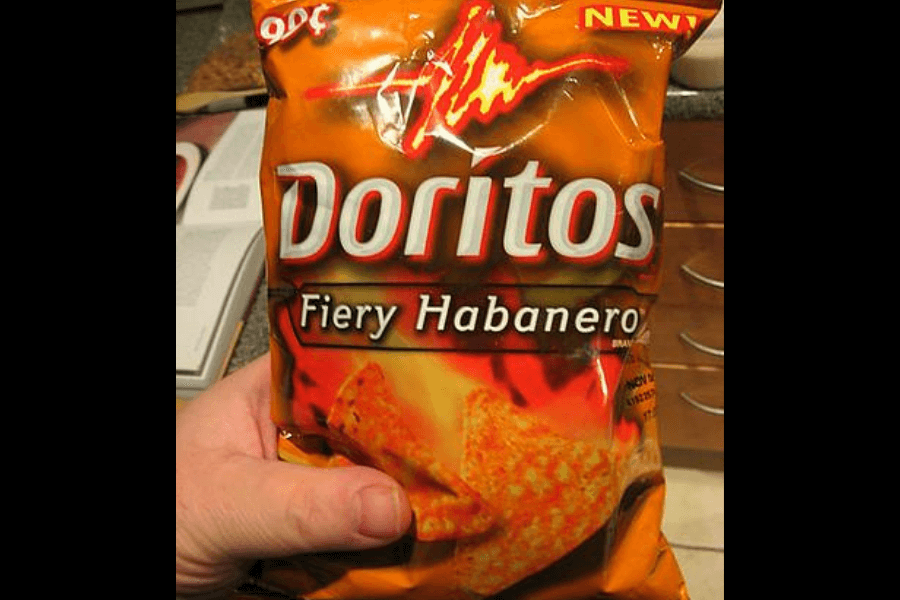 A bag of Doritos in the "Fiery Habanero" flavor, featuring a bold orange and red design. The packaging highlights the intense spiciness with flames and a chili pepper graphic, along with an image of seasoned triangular chips. The bag is labeled "New!" and is being held in a kitchen with drawers visible in the background.