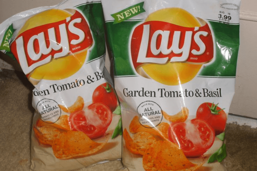 Two bags of Lay's chips in the "Garden Tomato & Basil" flavor, featuring a white and green design. The packaging showcases ripe tomatoes, fresh basil leaves, and crispy seasoned chips, emphasizing the natural and savory flavor. A "New!" label and "All Natural" badge are displayed prominently on the bags, which are placed on a carpeted surface.