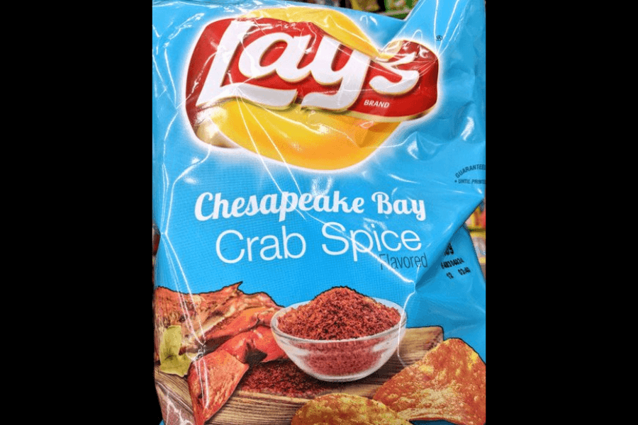 A bag of Lay's chips in the "Chesapeake Bay Crab Spice" flavor, featuring a bright blue design. The packaging showcases a bowl of crab seasoning along with images of crab legs and seasoned chips, highlighting the bold and savory flavor. The bag is displayed in a store environment with other products visible in the background.