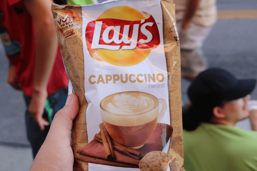 A bag of Lay's chips in the "Cappuccino" flavor, featuring a creamy cappuccino in a white mug with latte art and cinnamon sticks on the packaging. The bag includes a "Vote to Save Your Fave" promotion and is held by a hand outdoors with people visible in the background.