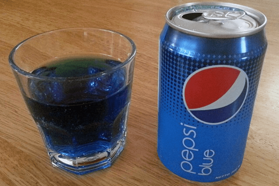 This image shows a can of "Pepsi Blue" next to a glass filled with the bright blue-colored soda. The can features a bold blue design with the Pepsi logo and text, placed on a wooden table.