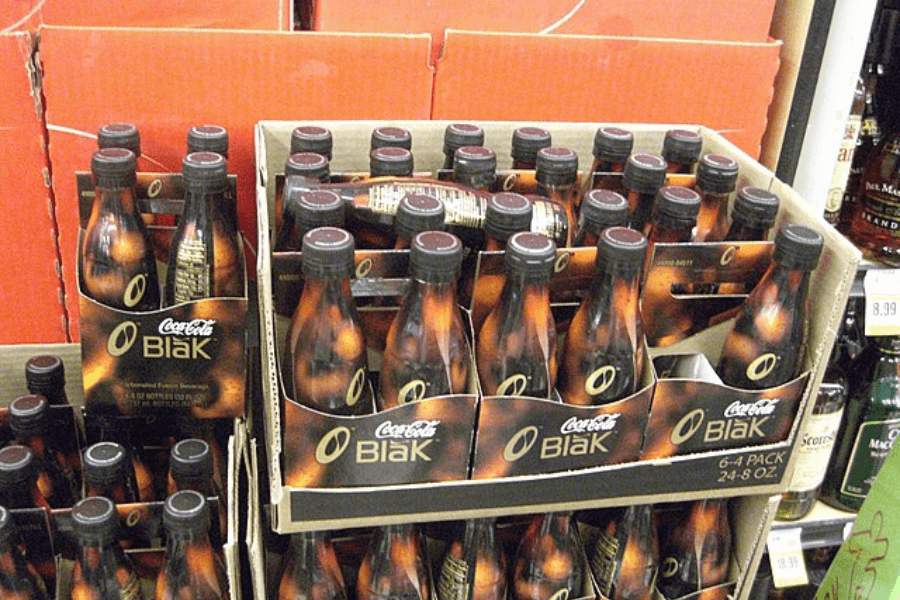 This image shows a store display of Coca-Cola BlāK, a coffee-infused soda. The bottles are packaged in sleek black and bronze-colored six-packs, arranged in cardboard boxes on a shelf.