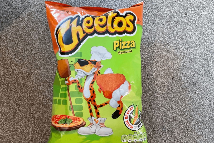 A bag of Cheetos in the "Pizza" flavor featuring a green and orange design. The packaging shows Chester Cheetah dressed as a chef, holding a pizza paddle and a large pizza-flavored Cheeto. A graphic highlights "Made of Corn" with an oven illustration in the corner. The bag is displayed on a carpeted floor.