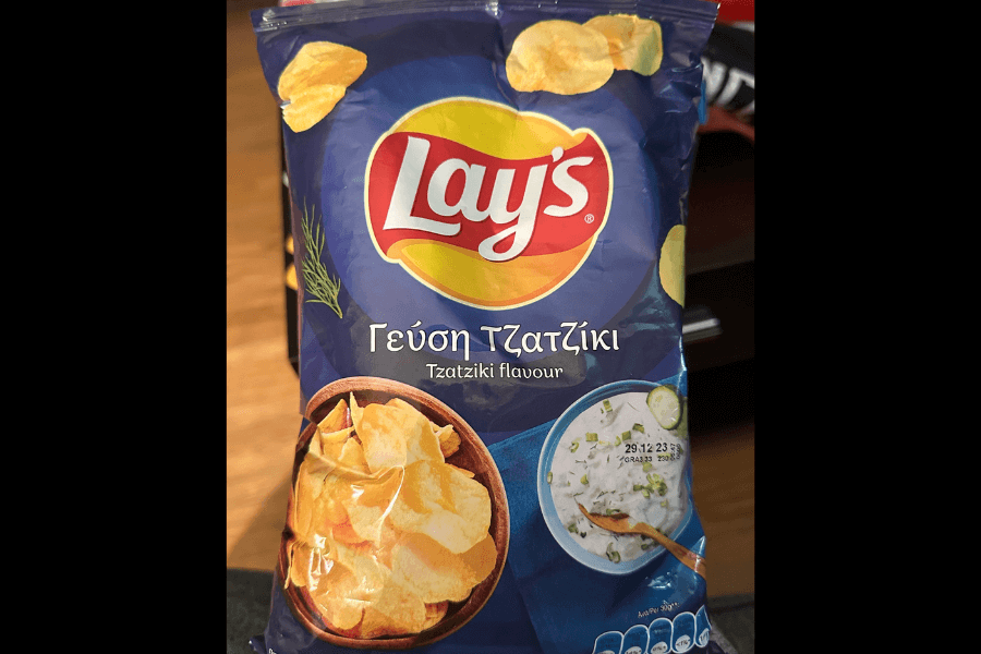 A bag of Lay's chips in the "Tzatziki" flavor, featuring a dark blue design. The packaging displays a bowl of crispy potato chips alongside a bowl of creamy tzatziki dip garnished with cucumber slices and dill. The label includes Greek text, emphasizing the flavor, and the bag is set against a warm indoor background.