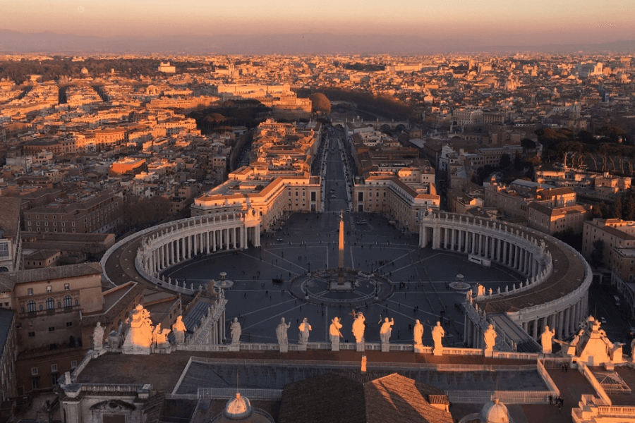 Aerial view of St. Peter’s Square in Vatican City during golden hour, showing the symmetrical colonnades encircling the plaza and the central obelisk, with the surrounding city of Rome bathed in warm sunset light. Statues of saints line the rooftop in the foreground.