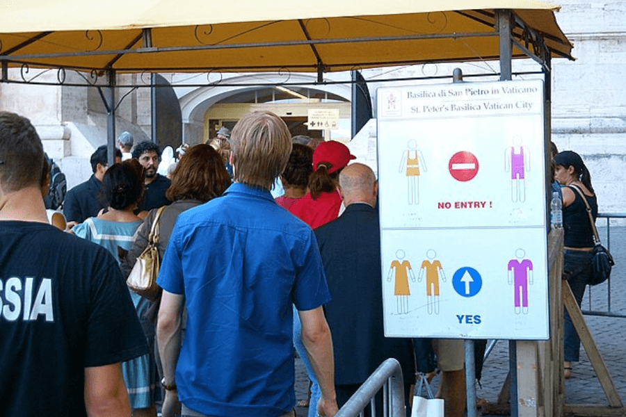 Crowd of visitors lining up outside St. Peter’s Basilica under a canopy, with a dress code sign prominently displayed that illustrates acceptable and unacceptable attire, indicating “No Entry” for sleeveless tops, shorts, and short skirts, and “Yes” for modest clothing covering shoulders and knees.