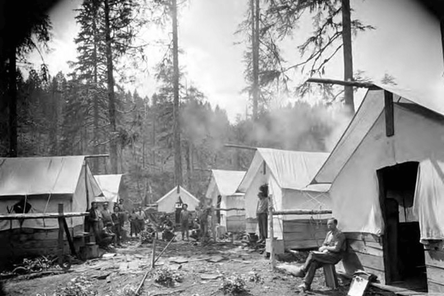 Black-and-white photo of a rustic outdoor camp with multiple canvas tents lined up in a forest clearing. Several people are gathered around a central area, some sitting and others standing, with smoke rising from fires, indicating cooking or warmth.