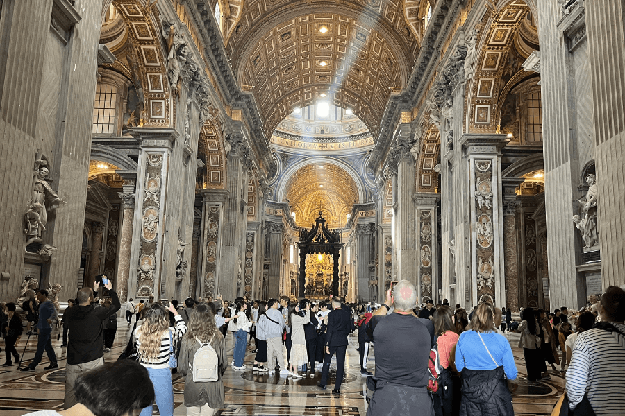 Interior of St. Peter’s Basilica filled with visitors admiring its grandeur, with intricately carved marble columns, soaring vaulted ceilings with golden coffering, and the ornate bronze Baldachin by Bernini above the papal altar in the distance.