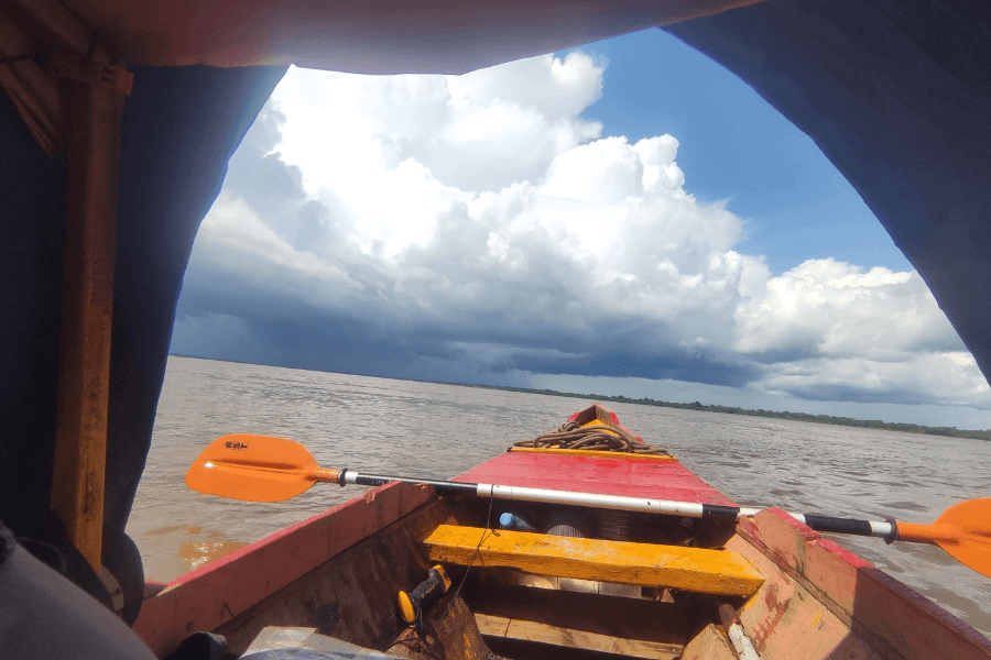 View from inside a small boat with a red and yellow wooden interior, looking out over a wide, muddy river under a dramatic sky with billowing white and dark storm clouds. Two orange paddles rest across the boat as it heads toward the horizon.