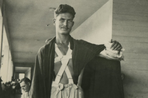 The True Story of the War Hero Who Fought Without Firing a Shot Black-and-white photo of a wounded soldier standing in a hospital or recovery ward, his chest wrapped in bandages forming an X pattern and his right arm in a heavy dressing. He wears loose pants and a blanket draped over his shoulders, with a calm but intense expression on his face.