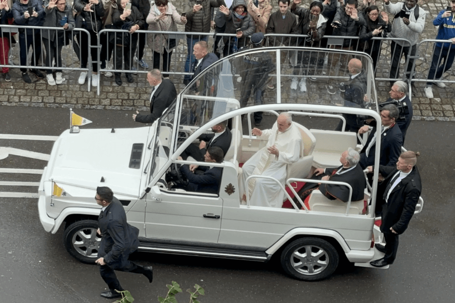 Pope Francis rides in a white popemobile with a glass enclosure, waving to crowds of people lined along barricades during a public procession; security personnel walk and run alongside the vehicle.