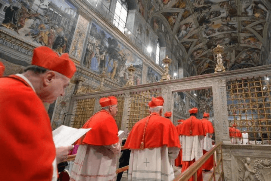 Catholic cardinals dressed in red vestments gather inside the Sistine Chapel beneath its iconic frescoed ceiling and walls, participating in a ceremonial event near a decorative wooden partition.