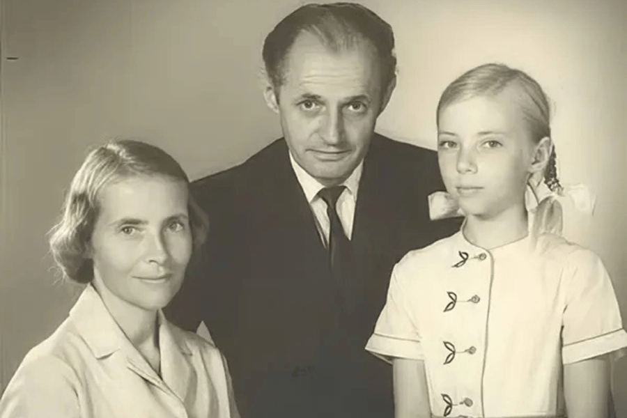 Sepia-toned studio portrait of a family, featuring a woman in a light blouse, a man in a dark suit and tie, and a young girl with braided blonde hair tied with large white bows, wearing a buttoned dress with leaf-shaped accents.