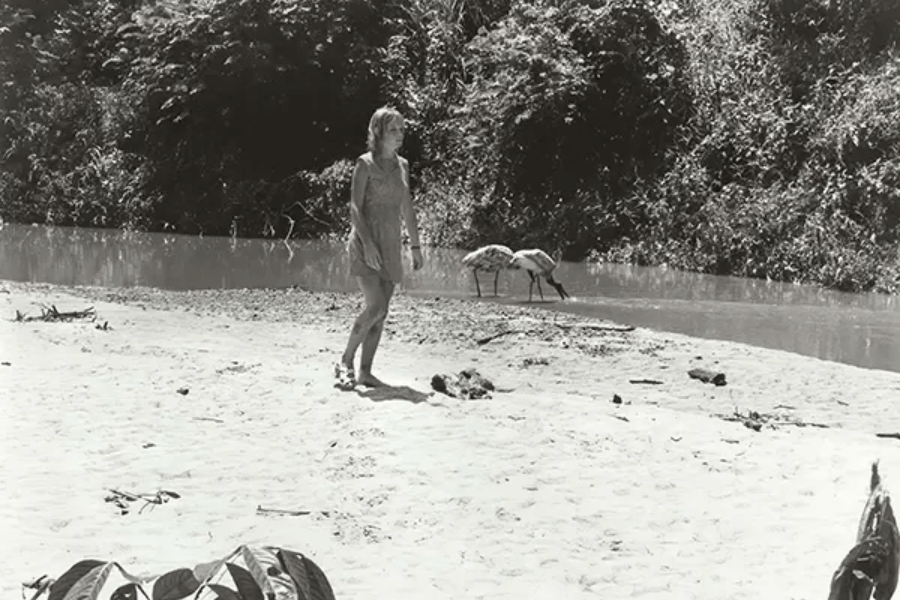 Black-and-white photo of a woman walking barefoot on a sandy riverbank, wearing a simple dress and looking toward the jungle. Two large birds are seen drinking from the river in the background, with dense forest foliage lining the opposite bank.